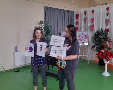 Sala. Fizjoterapeutki Martyna Józefczyk, Paulina Winczura pokazują seniorkom propozycje logo warsztatów kobiecych.