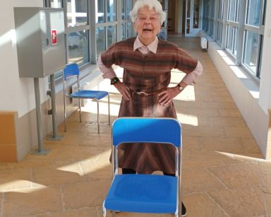 Korytarz. Seniorka stoi za krzesłem. Trzyma się pod boki. Śmieje się.