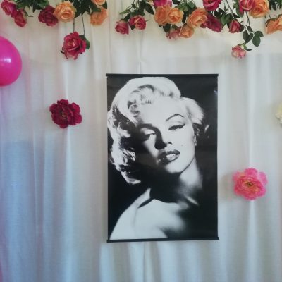 Dekoracja. Czarno-biały portret Marylin Monroe. Obok zawieszone sztuczne kwiaty, różowe balony.