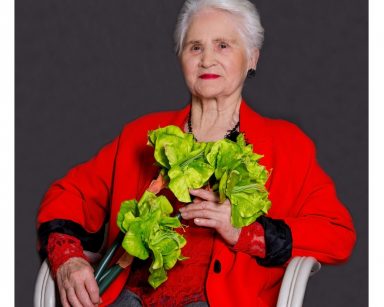 Seniorka pozuje do zdjęcia. Siedzi na krześle, uśmiecha się. Trzyma zielone kwiaty.