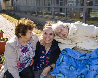 Słoneczny dzień. Patio. Dwie seniorki przytulają fizjoterapeutkę Martynę Józefczyk.