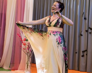Sala. Artystka Agnieszka Babicz śpiewa. W jednej ręce trzyma mikrofon, w drugiej róg kolorowej sukienki.