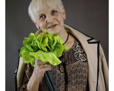 Seniorka pozuje do zdjęcia. Uśmiecha się, trzyma zielone kwiaty.