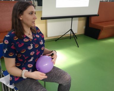 Sala gimnastyczna. Fizjoterapeutka Martyna Józefczyk siedzi na krześle, trzyma balon. Za nią ekran projektora.