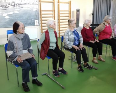 Sala gimnastyczna. Seniorki siedzą w rzędzie, na krzesłach.