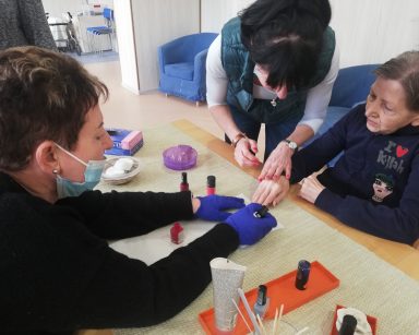 Świetlica. Kierowniczka Mariola Ludwicka maluje paznokcie seniorce. Obok nachyla się terapeutka Ania Rzepczyńska.