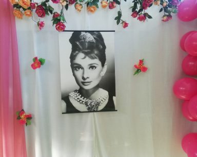 Dekoracja. Czarno-biały portret Audrey Hepburn. Obok zawieszone sztuczne kwiaty, różowe balony.