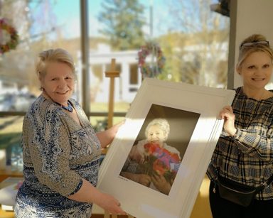 Ogród zimowy. Terapeutka Beata Gadomska i seniorka trzymają oprawiony w białą ramę portret seniorki.