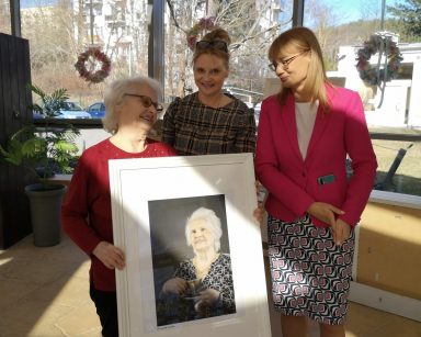 Ogród zimowy. Roześmiana seniorka trzyma swój portret. Obok dyrektorka Agnieszka Cysewska, terapeutka Beata Gadomska.