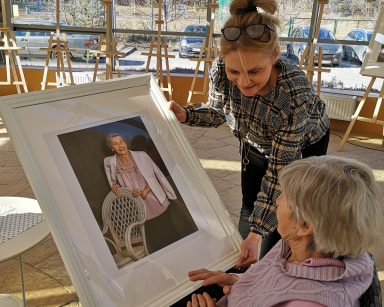 Ogród zimowy. Terapeutka Beata Gadomska, trzyma zdjęcie w ramie. Nachyla się nad seniorką. Seniorka ogląda swój portret.