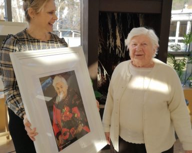 Ogród zimowy. Terapeutka Beata Gadomska i seniorka stoją obok siebie. Śmieją się. Terapeutka trzyma portret seniorki.