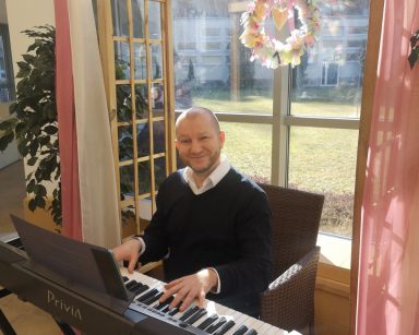 Słoneczny dzień. Ogród zimowy. Na szybie kolorowy wieniec. Przy pianinie siedzi kierownik Arkadiusz Wanat. Uśmiecha się, gra.