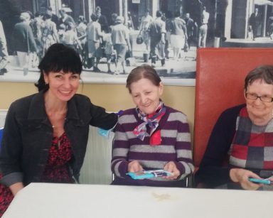 Sala. Przy stole terapeutka Ania Rzepczyńska i dwie seniorki. Każda z seniorek trzyma tabliczkę czekolady.