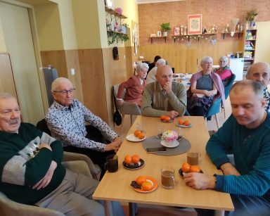 Sala. Seniorzy siedzą przy stołach. Na blatach szklanki z kawą, talerzyki z piernikami i mandarynkami.