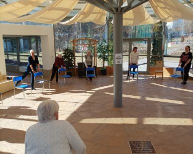 Słoneczny dzień. Ogród zimowy. Terapeutki i seniorzy stoją w kręgu. Ćwiczą przy krzesłach.