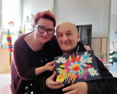 Świetlica. Koordynatorka Edyta Życzyńska i senior pokazują obrazek z kwiatów z kolorowego papieru.