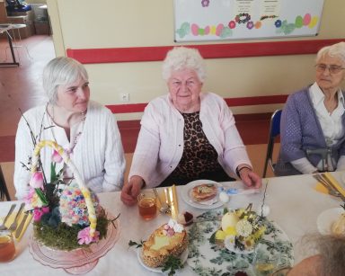 Przy stole trzy seniorki. Stół udekorowany wielkanocnymi ozdobami. Na talerzach mazurki.