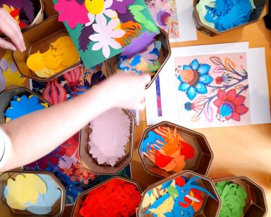 Widok z góry. Na stole pudełka z kolorowymi kwiatami z wycinanek. Ręce osoby układającej obrazek z papierowych kwiatów.