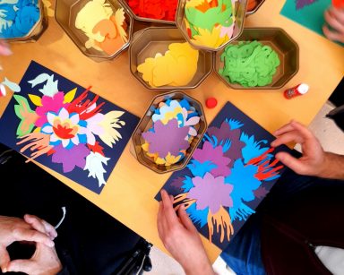 Widok z góry. Na stole pudełka z kolorowymi kwiatami z wycinanek. Ręce osób układających obrazki z papierowych kwiatów.