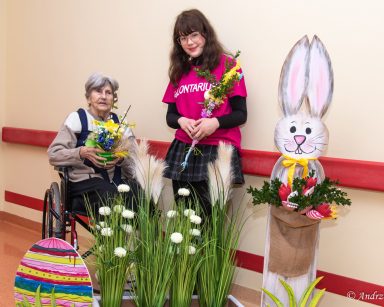 Przy wielkanocnych dekoracjach seniorka i wolontariuszka. Wolontariuszka trzyma palmę z bukszpanu, seniorka stroik.