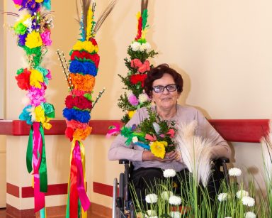 Seniorka pozuje do zdjęcia przy wielkanocnych dekoracjach. Uśmiecha się. Trzyma palmę z bukszpanem.