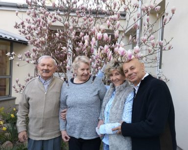 Słoneczny dzień. Przy magnolii z różowymi kwiatami pozują dwie seniorki i dwóch seniorów. Obejmują się i uśmiechają.