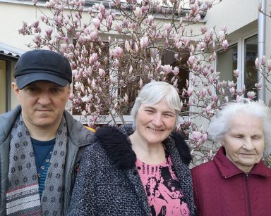 Słoneczny dzień. Przy magnolii z różowymi kwiatami pozuje troje seniorów. Uśmiechają się.