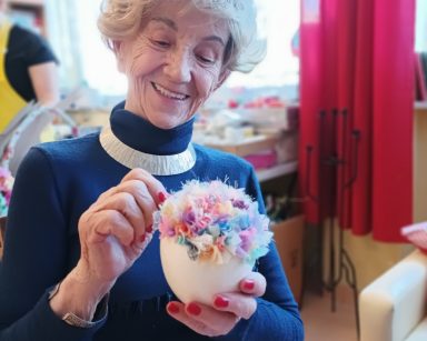 Sala. Seniorka dekoruje jajo ze styropianu kolorowym materiałem. Uśmiecha się.
