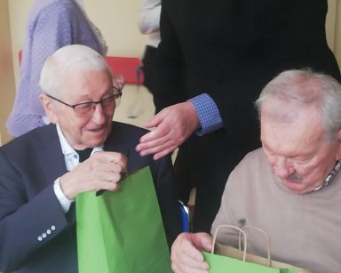 Dwóch seniorów przy stole. Trzymają zielone torebki z upominkami. Obok stoi ksiądz Krzysztof Rybka. Śmieje się.