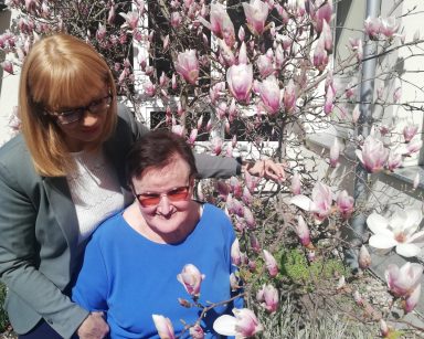Ogród. Przy magnolii z różowymi kwiatami seniorka i dyrektorka Agnieszka Cysewska. Uśmiechają się.