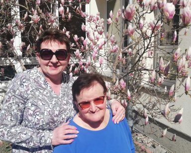 Ogród. Przy magnolii z różowymi kwiatami seniorka i kierowniczka Mariola Ludwicka. Uśmiechają się.