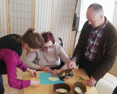 Świetlica. Wolontariuszka z Ukrainy Mariczka i Vłodek Ivanets pomagają seniorce. Wspólnie układają kwiaty z papieru.