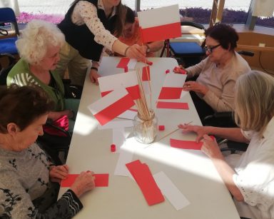 Ogród zimowy. Przy stole seniorki i wolontariuszka Mariczka. Robią biało-czerwone flagi z papieru.