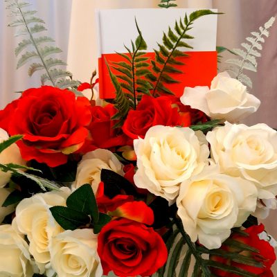 Bukiet z białych i czerwonych róż. Jest ozdobiony liśćmi paproci i flagą Polski.
