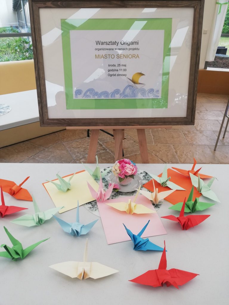 Ogród zimowy. Na sztalugach plakat zapraszający na zajęcia z origami. Na stole ptaki złożone z papieru.