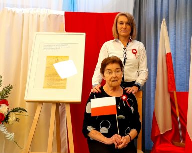 Zdjęcie portretowe. Seniorka siedzi na krześle. Trzyma flagę Polski. Za nią stoi zastępca dyrektora Ilona Gajewska.