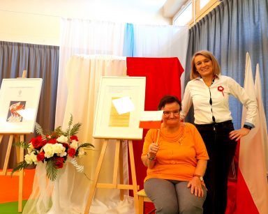 Zdjęcie portretowe. Seniorka siedzi na krześle. Trzyma flagę Polski. Obok zastępca dyrektora Ilona Gajewska. Śmieją się.