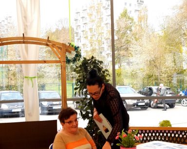 Ogród zimowy. Przy stole siedzi seniorka. Obok nachyla się terapeutka Magdalena Poraj-Górska.