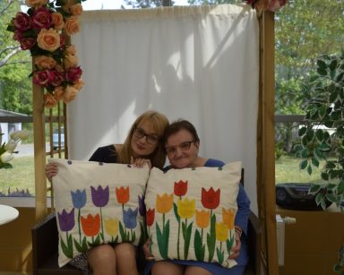Ogród zimowy. Pergola. Dyrektor Agnieszka Cysewska i seniorka siedzą obok siebie. Trzymają poduszki z wyszytymi tulipanami.