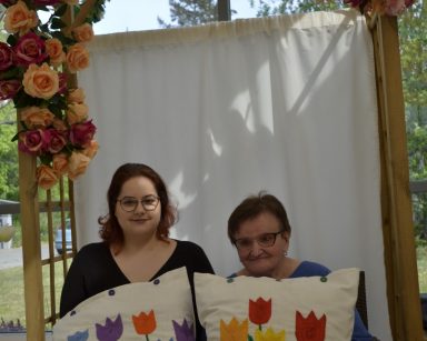 Ogród zimowy. Pergola. Psycholog i seniorka siedzą obok siebie. Trzymają poduszki z wyszytymi tulipanami.
