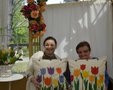Ogród zimowy. Pergola. Terapeutka Gosia Jancelewicz i seniorka siedzą obok siebie. Trzymają poduszki z wyszytymi tulipanami.