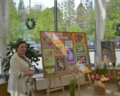 Ogród zimowy. Terapeutka Gosia Jancelewicz stoi przy tablicy z obrazkami. Prezentuje wystawę.