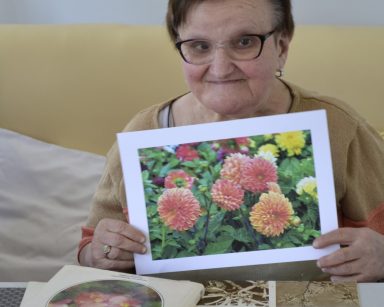 Pokój. Seniorka siedzi przy stole. Uśmiecha się. Pokazuje zdjęcie kolorowych kwiatów.