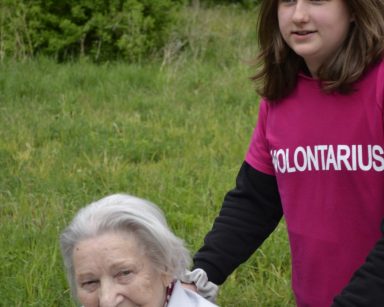 Spacer na świeżym powietrzu. Wolontariuszka pomaga seniorce na wózku inwalidzkim.