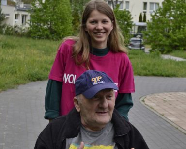 Na świeżym powietrzu spaceruje wolontariuszka z seniorem na wózku. W tle budynek Domu Pomocy Społecznej w Sopocie.