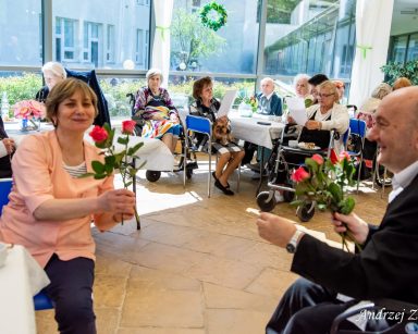 Ogród zimowy. Senior daje opiekunce różę. Dalej przy stolikach siedzą pracownicy i seniorzy.
