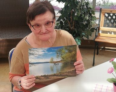 Ogród zimowy. Seniorka siedzi przy stole. Trzyma z zdjęcie. Na zdjęciu jezioro i drzewa.
