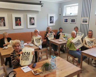 Jasna sala. Przy stołach siedzą kobiety. Każda trzyma bawełnianą siatkę z napisem Dom Pomocy Społecznej w Sopocie.