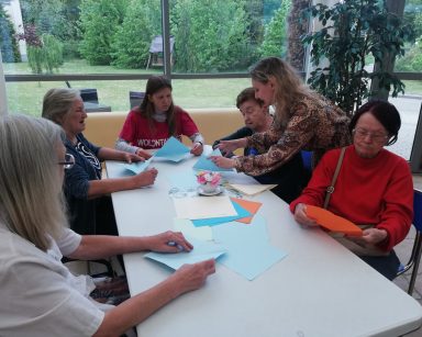 Ogród zimowy. Warsztaty origami. Seniorzy, wolontariuszka i pracownicy przy stole. Składają figury z papieru.
