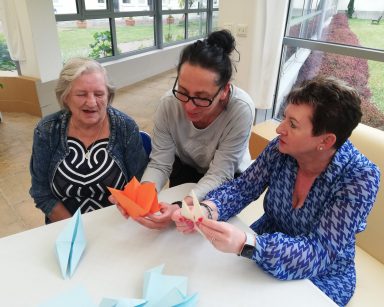 Ogród zimowy. Kierowniczka Mariola Ludwicka, terapeutka Magdalena Poraj-Górska i seniorka składają figury techniką origami.